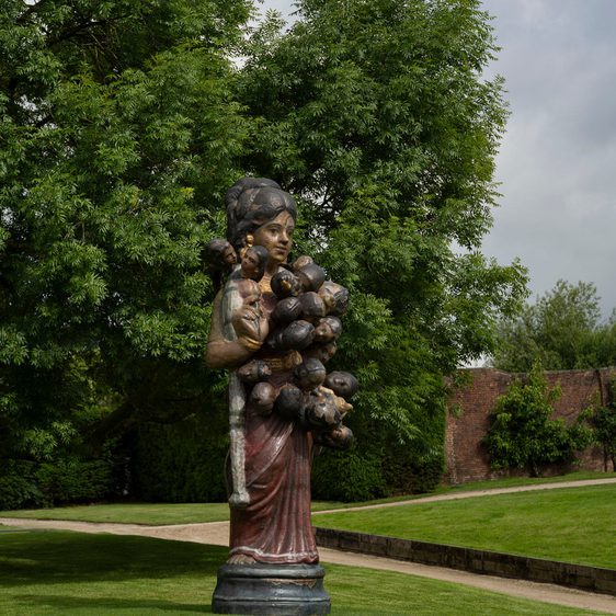 A sculpture outside on grass depicting a woman with lots of heads attached to her body