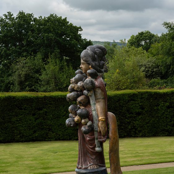 A sculpture outside on grass depicting a woman with lots of heads attached to her body