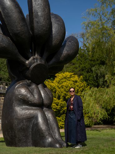 A woman wearing a navy coat and trousers, standing next to a large black seated figure with bananas on it's head.