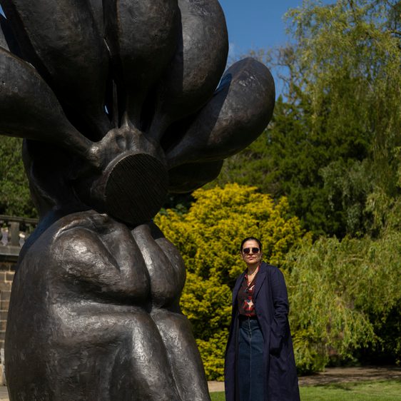 A woman wearing a navy coat and trousers, standing next to a large black seated figure with bananas on it's head.