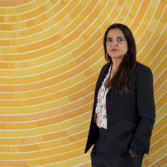 A woman wearing a black suit standing in front of a yellow spiral wall-based artwork.