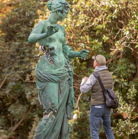 A man taking a photograph of a giant bronze sculpture of a naked woman, with eroded bronze crystallised sections.
