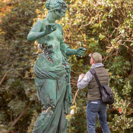 A man taking a photograph of a giant bronze sculpture of a naked woman, with eroded bronze crystallised sections.