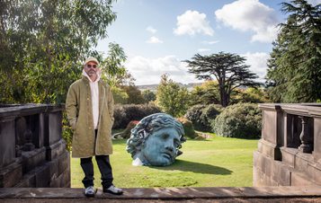 A man wearing a tan jacket and baseball cap, standing in front of a sculpture of a woman's head with eroded crystal sections.