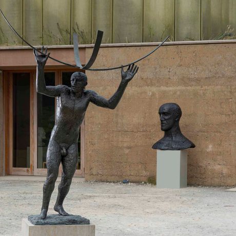 A bronze sculpture of a naked male figure holding curved bars above his head, displayed outdoors. A sculpture of a head displayed on a plinth is behind it, next to a low stone building.