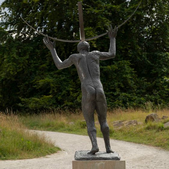 The rear of a bronze sculpture of a naked male figure holding curved bars above his head, displayed outdoors.