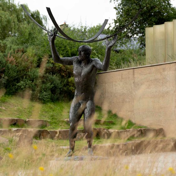 A bronze sculpture of a naked male figure holding curved bars above his head, displayed outdoors.
