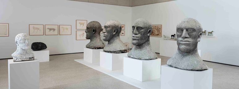 Four large head sculptures displayed on plinths in a row inside a gallery space.