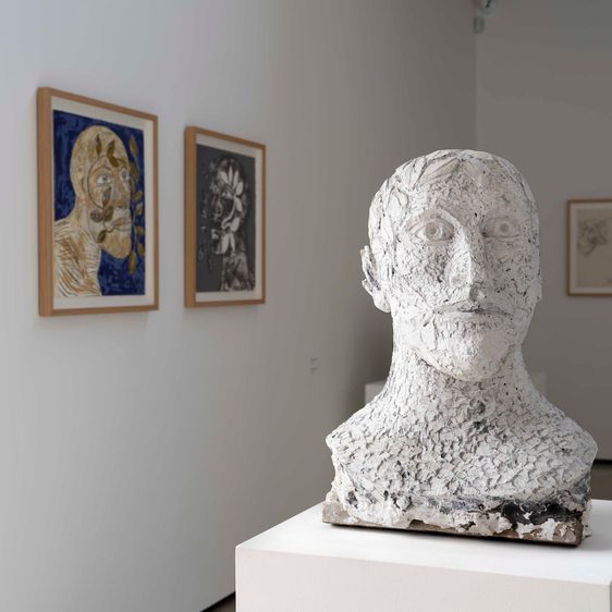A sculpture of a human head displayed on a plinth, with two framed prints on the wall behind it.