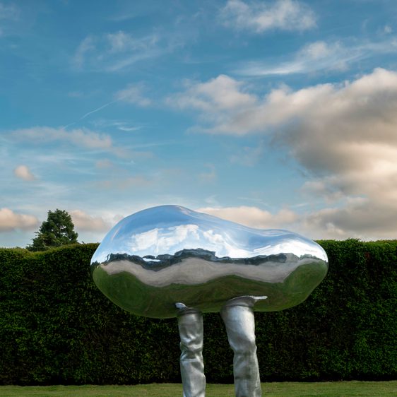 A silver pebble with legs sculpture, stood on grass underneath a blue sky with grey clouds