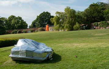 A silver squashed car on the lawn and a orange hot water bottle in the distance
