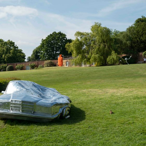 A silver squashed car on the lawn and a orange hot water bottle in the distance