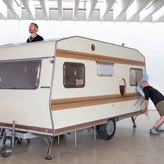 A caravan with a man poking his head out of the top and another man leaning on a blue chair attached to the side of the caravan