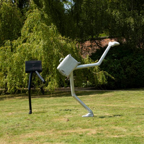 A silver suitcase sculpture and black briefcase sculpture both with legs on a lawn.