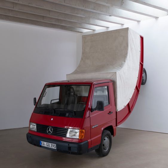 A bent red truck against a wall inside a gallery
