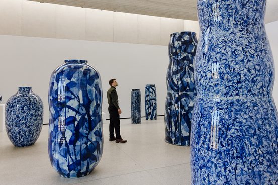A man wearing dark clothing, standing in a gallery space filled with large ceramic pots which tower above him. The ceramics are decorated with blue and white patterns.