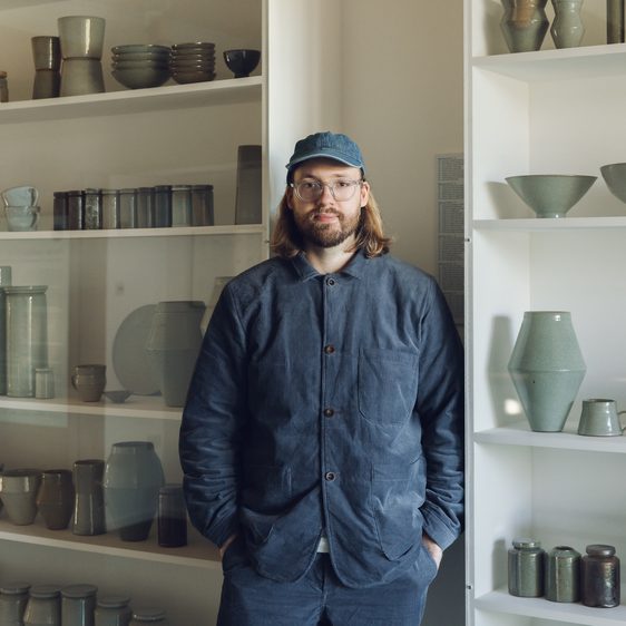 Man with shoulder length hair wearing glasses and a cap standing in front of shelves packed with ceramic vessels