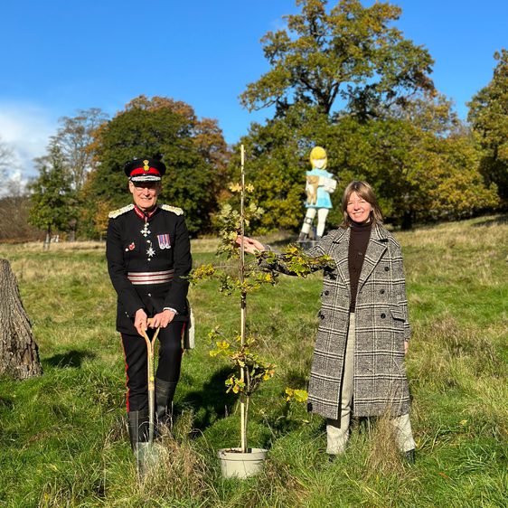 Man and woman in a grassy field, stood next to a sapling oak tree