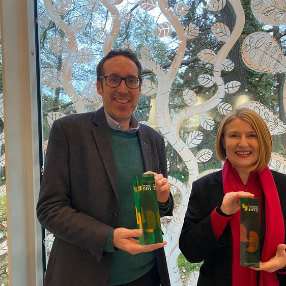 Man wearing a suit and glasses holding a glass award next to a woman wearing a pink scarf holding a glass award.