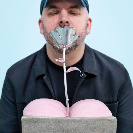 Man smelling a silver rose growing between sculpture of two pink bottom cheeks
