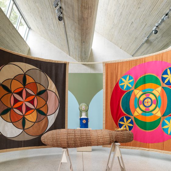 Indoor gallery space with two hanging tapestries and a woven basket coffin