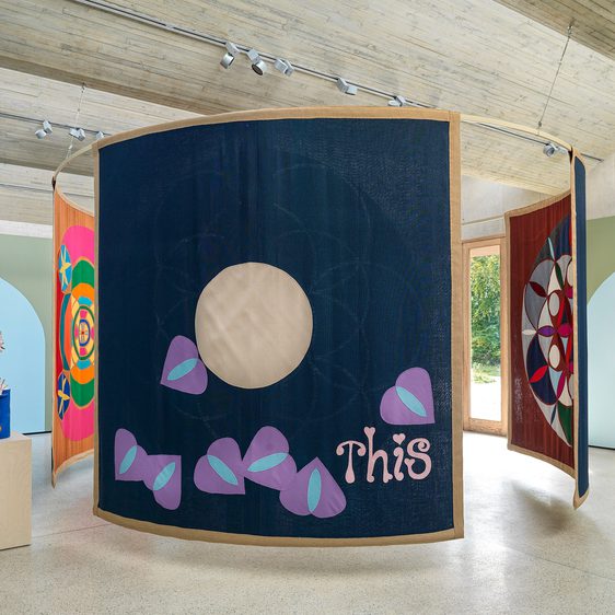 Inside gallery space with three tapestries hanging from the ceiling in the centre of the room in a circle, with several large statues on plinths