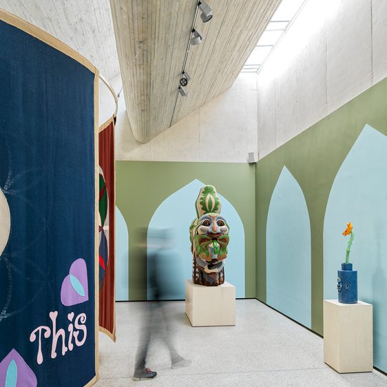 Indoor gallery space with a tapestry hanging from the wall and two large sculptures on plinths