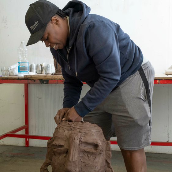 A black woman wearing shorts and a sweatshirt, working on a clay head sculpture