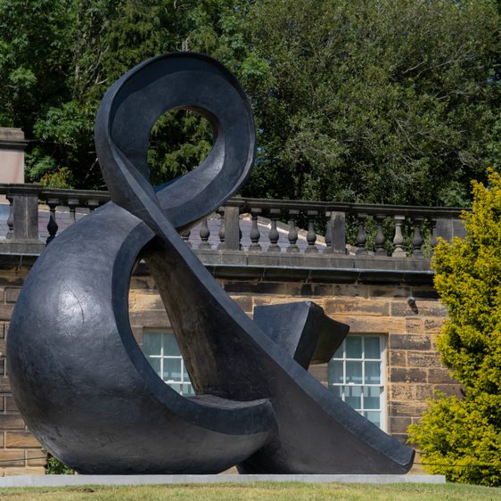 A large, abstract black sculpture resembling an ampersand sits on a grassy area, surrounded by trees. In the background, a stone balustrade adds architectural interest.