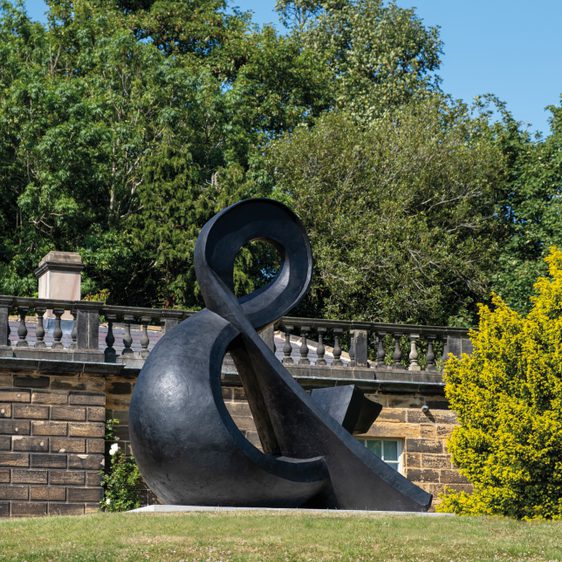 A large, abstract black sculpture resembling an ampersand sits on a grassy area, surrounded by trees. In the background, a stone balustrade adds architectural interest against a clear blue sky.