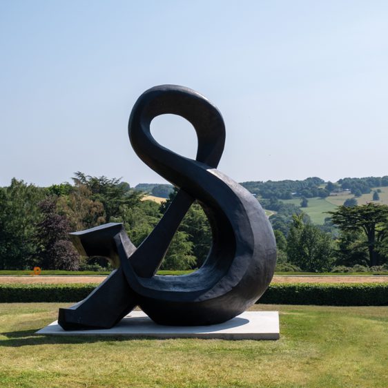 A large, abstract black sculpture resembling an ampersand stands on a grassy area, with rolling green hills and trees in the background under a clear blue sky. The sculpture's smooth curves contrast with the natural landscape.