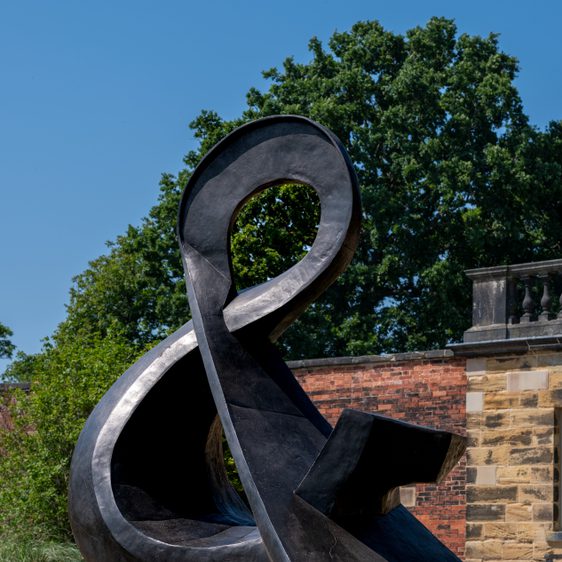 A large, abstract black sculpture resembling an ampersand sits on a grassy area, surrounded by trees. In the background, a stone balustrade adds architectural interest against a clear blue sky.