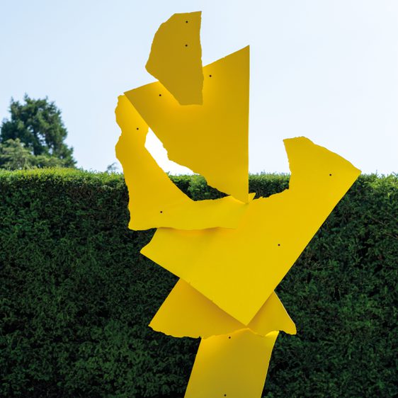 A large, abstract yellow sculpture stands prominently on a grassy area, surrounded by neatly trimmed hedges. The sculpture features dynamic, angular shapes that suggest movement and energy.