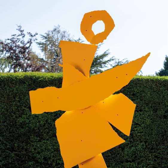 A large abstract sculpture made of bright orange metal, featuring geometric shapes and a circular element at the top. It stands on a black base, surrounded by a neatly trimmed green hedge and a clear blue sky in the background.