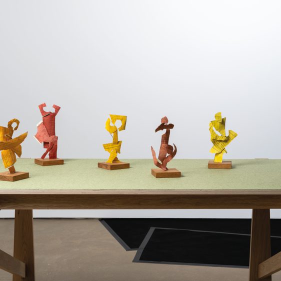 A wooden table with a green surface displays six abstract sculptures in vibrant colours, including red, yellow, and orange. The figures are dynamic and stylised, showcasing various poses, set against a plain white wall.