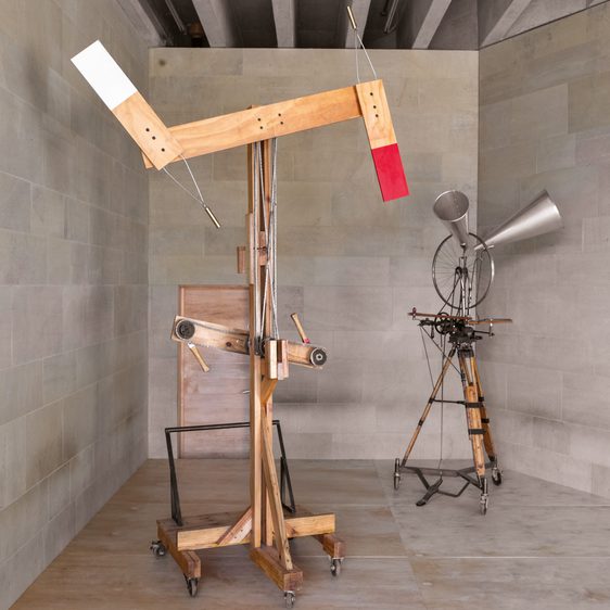 A wooden kinetic sculpture stands in a minimalist gallery, featuring a large arm with red and white ends. Beside it, another intricate apparatus with a horn-like structure and various mechanical components is displayed against a stone wall.