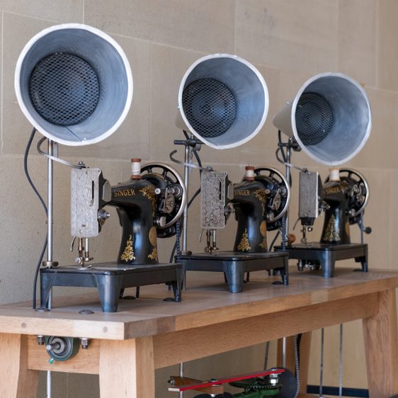 Three vintage Singer sewing machines are displayed on a wooden table. Each machine features a large, round speaker attached to the top, creating a unique blend of old and new technology against a plain stone wall backdrop.