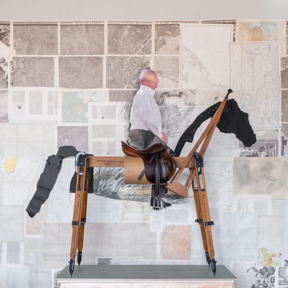 A man sits atop a large, wooden horse sculpture, which is mounted on a crate. Behind him, a wall features a collage of maps and drawings, creating a striking contrast with the sculpture's rustic appearance. The setting is bright and modern.