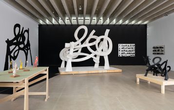 An art gallery featuring a large white abstract sculpture with looping forms at its centre. Surrounding it are smaller sculptures in various colours and styles displayed on wooden tables, against a contrasting black wall.