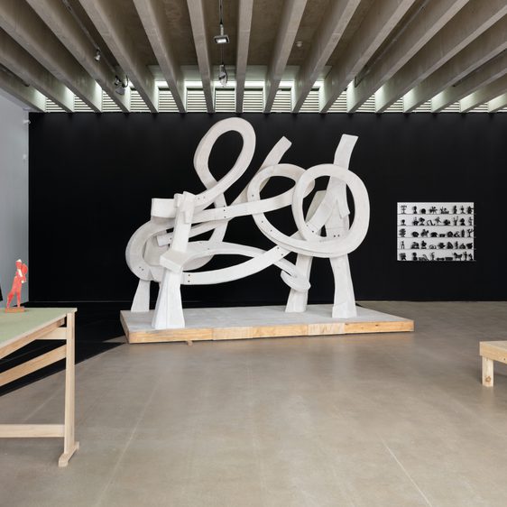 An art gallery featuring a large white abstract sculpture with looping forms at its centre. Surrounding it are smaller sculptures in various colours and styles displayed on wooden tables, against a contrasting black wall.