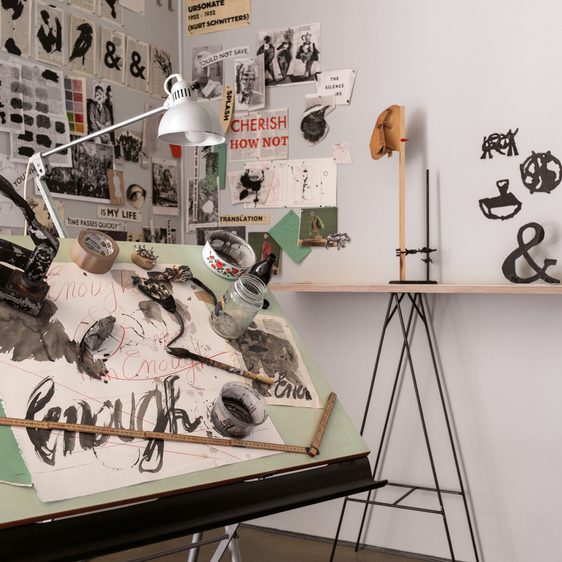 A cluttered art studio featuring a large easel with mixed media artworks, including sketches and collages. Various tools and materials are scattered across the surface, alongside a lamp. The wall displays an array of black-and-white posters and notes.