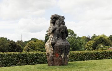 A textured sculpture of a human figure with two faces, stands on a grassy lawn. Trees and hedges can be seen behind it.