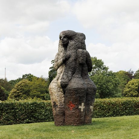 A textured sculpture of a human figure with two faces, stands on a grassy lawn. Trees and hedges can be seen behind it.