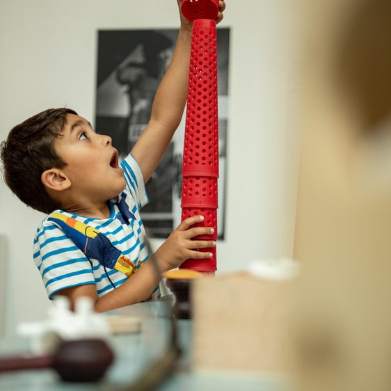 An excited child using red plastic cones to create a sculpture. They are in an indoor space with black and white photographs on the walls.