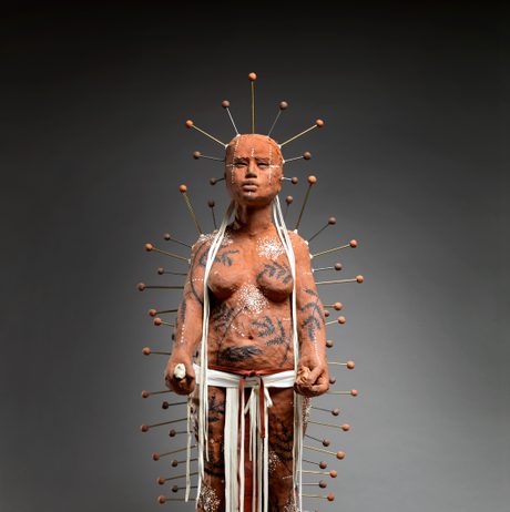A life-sized sculpture of a figure covered in intricate body art and clay, standing confidently. The figure is adorned with numerous pins radiating from the head, and white ribbons hang from the waist, set against a dark background.