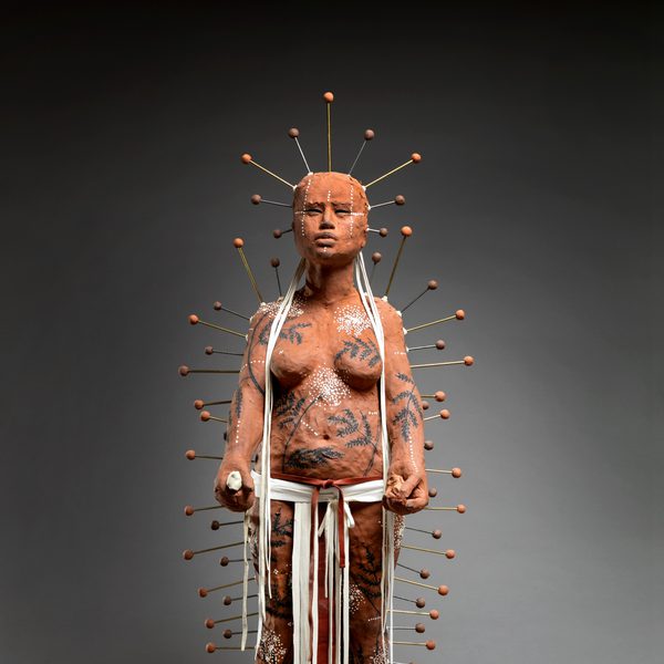 A life-sized sculpture of a figure covered in intricate body art and clay, standing confidently. The figure is adorned with numerous pins radiating from the head, and white ribbons hang from the waist, set against a dark background.
