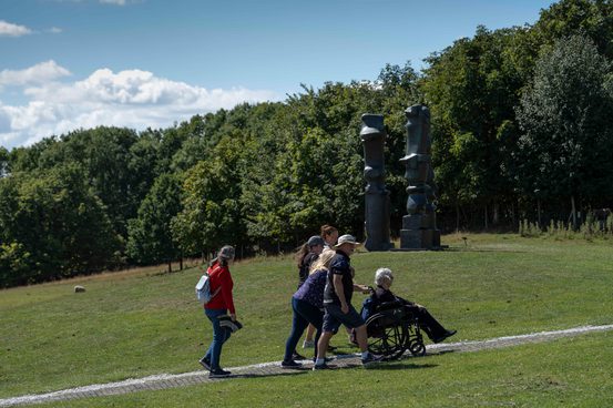A group of people, including a wheelchair user, walking along an outdoor pathway. Three tall bronze sculptures are in the background.