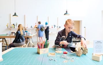 A young boy focuses on assembling a wooden model at a craft table, surrounded by tools and materials. In the background, adults browse a display of creative projects. The space is bright and inviting, with pendant lights and a minimalist design.