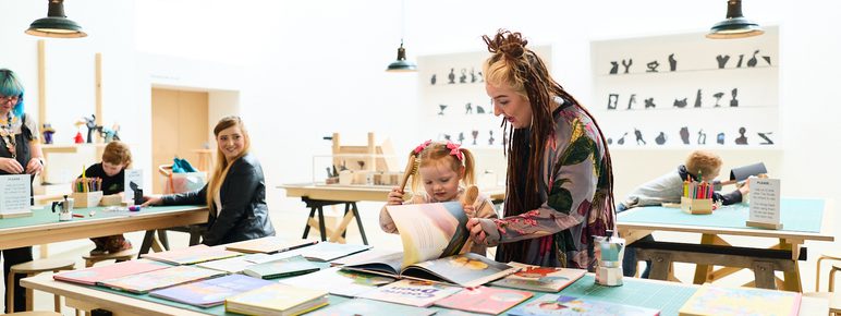 A bright, spacious art studio features several wooden tables covered with colourful books and art supplies. A woman with long hair helps a young girl with a book, while other participants engage in creative activities in the background.