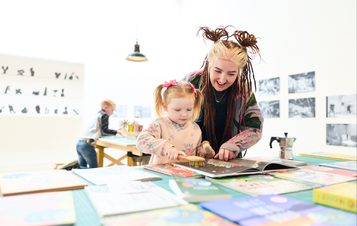 A young girl and a woman with styled hair are engaged in a creative activity at a table covered with colourful books and art supplies. The girl is using a brush on a book, while the woman smiles encouragingly. In the background, a boy is working at another table.